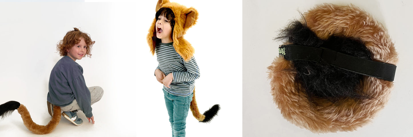 A child in a Lazy Lion costume by TellTails crouches playfully at a festival, while another in stripes laughs and holds a plush tail. The close-up shows the tails black adjustable strap on the right.