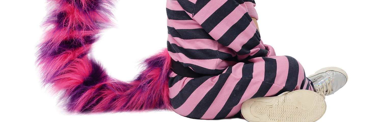 A child in The Crazy Cat pajamas by TellTails, featuring pink and black stripes with a furry tail, sits showcasing their sneakers. The playful fancy dress adds charm against the white background.