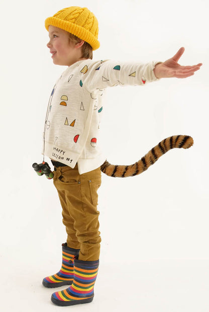 A child joyfully embodies the spirit of cosplay. They're wearing TellTails The Tiger tail, along with a colourful shirt, brown pants, striped rubber boots against a white backdrop.