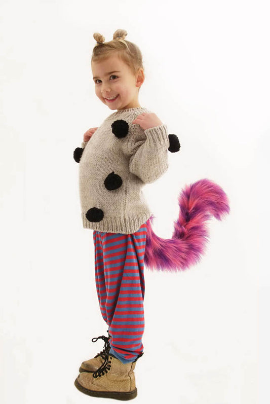 A young child playfully poses in The Crazy Cat tail by TellTails, featuring a sweater with black pompoms, vibrant purple and pink striped pants, brown boots, and a purple furry tail. Two small buns styled in their hair add flair to this delightful cosplay against the plain white background.