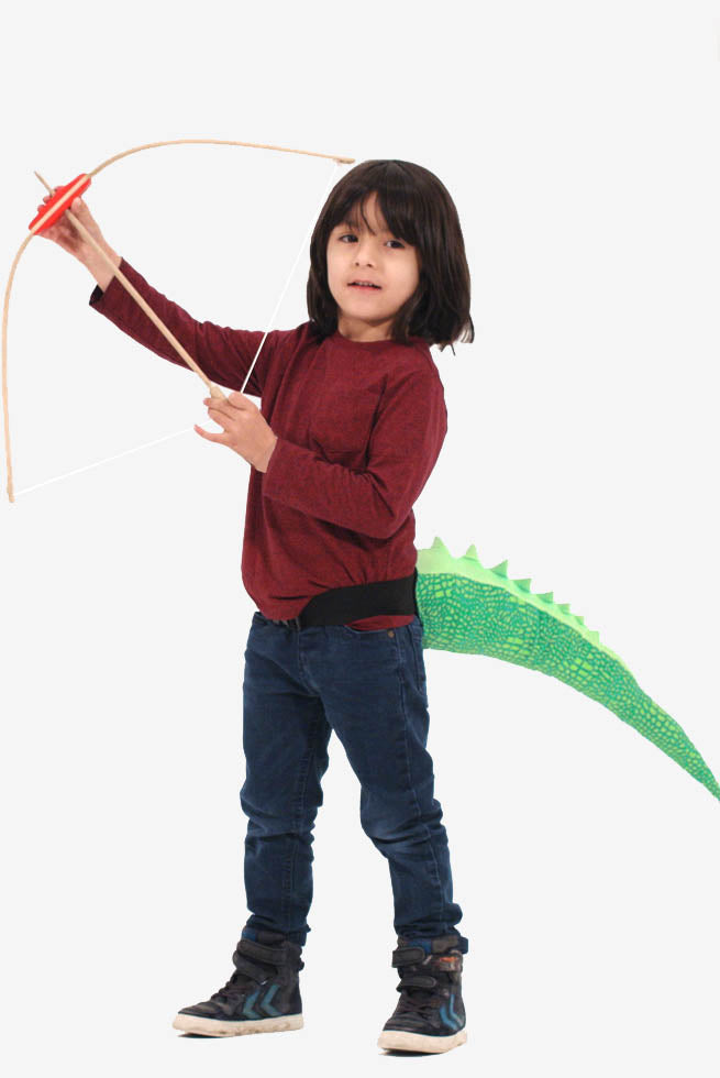 A kid aged 5 years old shooting a bow and arrow and wearing a dinosaur tail that's green and scaley that buckles on with a waistband that fits all ages. The tail is the perfect accessory for dino themed parties, jurassic world fans, a day out with our prehistoric friends at the Natural History Museum and any crocodile or cameleon charters for World Book Week like Gecko from PJ masks or how to train your dragon. But most of all like pouncing around music festivals, swooshing your tail as you go.
